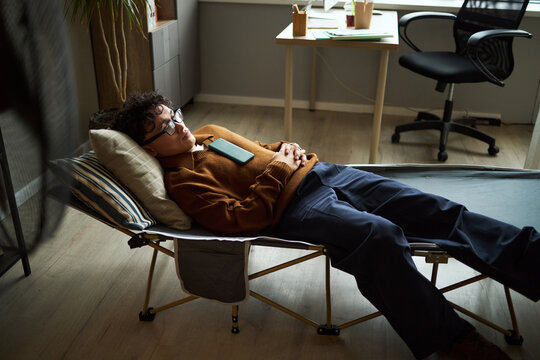 Young adult Asian man lying on office cot resting with smartphone on chest, wearing glasses, relaxing during workday in modern workspace, desk and chair visible in background