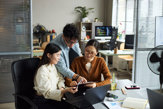 Young Asian businessmen and businesswomen collaborating in modern office, two young Asian women sitting at desk using digital tablet while young Asian man standing beside them pointing at screen