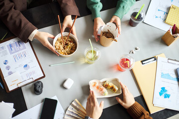 Young Asian businessmen and businesswomen sitting at office table eating lunch with chopsticks, discussing documents and charts, hands visible holding food containers and drinks during work