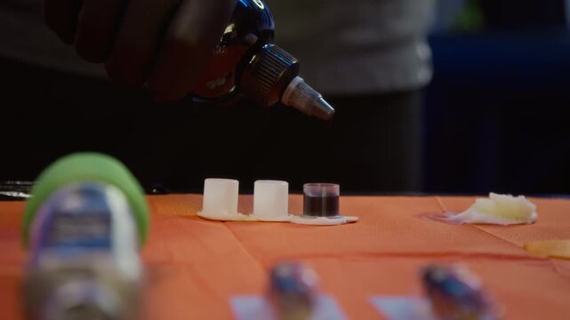 Cropped close up shot of hand of unrecognizable tattoo artist in black sterile gloves pouring ink in separate ink caps, preparing pigment for tattoo process in studio