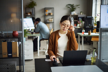 Young Asian woman working at desk using laptop and mouse, adjusting glasses, focused on screen, young Asian man sitting in background talking on phone in modern office workspace
