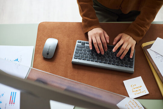 Young Asian woman working at office desk typing on wireless keyboard with hands visible, surrounded by business documents and notes, engaging in professional business tasks