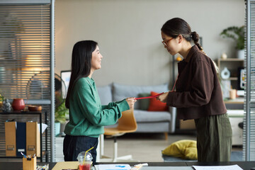 Young adult Asian businesswoman handing document to young adult Asian businesswoman in modern office setting, both standing and interacting over desk with paperwork visible