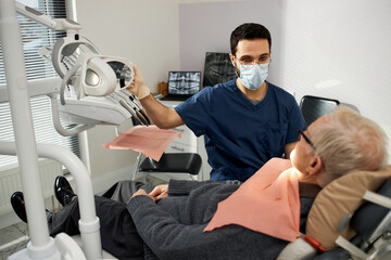 Middle Eastern male dentist wearing mask examining senior Caucasian man reclining in dental chair during dental appointment in modern dental office, dentist holding dental equipment