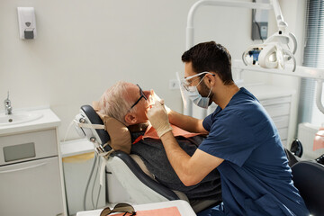 Senior Caucasian man sitting in dental chair receiving oral examination from young adult male dentist wearing protective eyewear and gloves in modern dental office