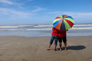 Obraz premium A Young couple on a romantic holiday holding a colorful umbrella on a sunny day immersed in watching the picturesque beach, ocean water waves and the sky in Kerala, India.