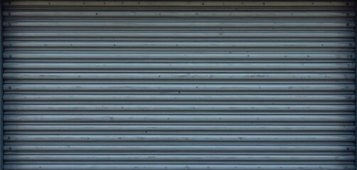 Grey metal roller shutter door creates industrial background. Striped steel surface of closed garage gate forms abstract texture. Weathered iron panel horizontal line pattern, simple urban wallpaper.