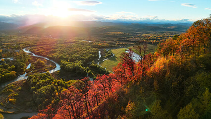 Golden sunset bathes a winding river valley and colorful autumn forest, viewed from above. Lens flare glows over distant mountains and rolling hills, creating a warm, cinematic panorama. At dusk.