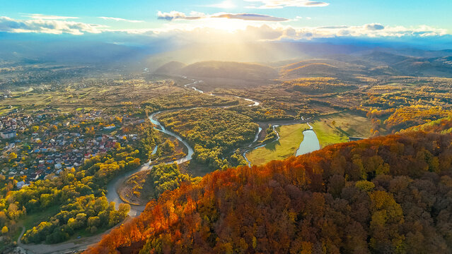 Sweeping aerial panorama of a winding river through autumn forest and town, sunbeams breaking through clouds over distant mountains, revealing the golden valley and scenic countryside from above. - Powered by Adobe