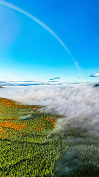 Aerial panorama of a foggy mountain forest partly submerged beneath a rolling sea of clouds. A faint rainbow curves across the blue sky, adding wonder to the autumn landscape and endless horizon.
