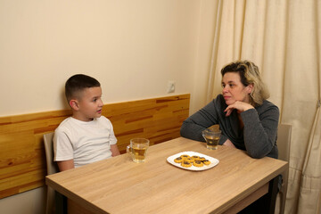 Family moment with mother and son sitting at table, drinking tea, eating cookies, and enjoying warm conversation together indoors.