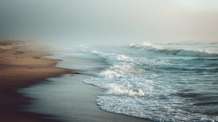 Gentle waves hit the shore of a sandy beach early in the morning. A thick fog covers the landscape creating a serene and tranquil atmosphere as the sun begins to rise.