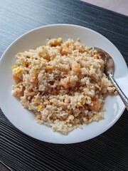 A white plate containing shrimp fried rice mixed with egg served with a spoon on a dark table.