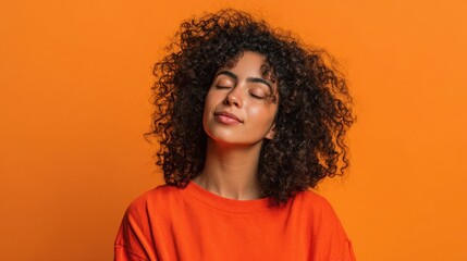 A young woman with curly hair stands against a vivid orange backdrop. She closes her eyes and smiles softly showcasing a moment of tranquility and happiness.