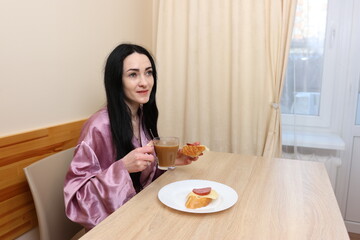 A woman is having breakfast with coffee and a sandwich while sitting at the table in the kitchen.