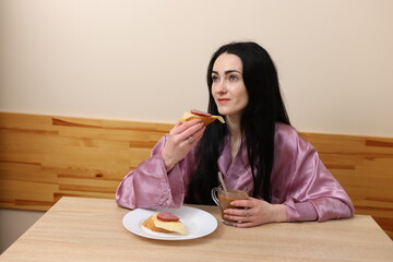 A woman is having breakfast with coffee and a sandwich while sitting at the table in the kitchen.