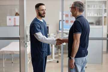 Male dermatologist greeting middle aged Caucasian man with handshake in medical office, both...