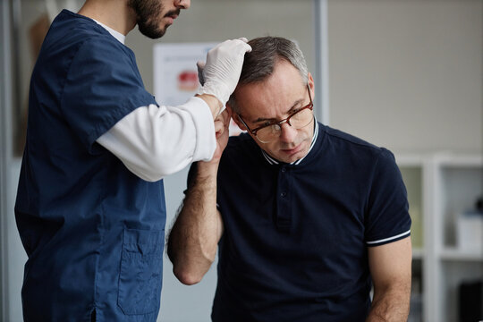 Middle aged Caucasian man receiving scalp examination from male healthcare professional wearing gloves during dermatology appointment, patient tilting head forward for inspection
