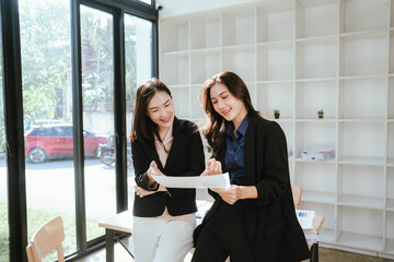 Two businesswomen work together at a laptop, smiling as they collaborate in an office with stacks of documents on the desk.