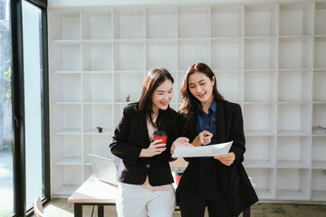 Two businesswomen work together at a laptop, smiling as they collaborate in an office with stacks of documents on the desk.