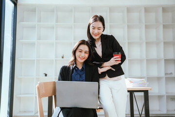 Two businesswomen work together at a laptop, smiling as they collaborate in an office with stacks of documents on the desk.