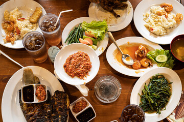 view from above, eating together, with a menu of rice, grilled chicken, chili sauce, fresh vegetables, crispy flour squid, iced orange tea, typical Indonesian food, on a wooden table