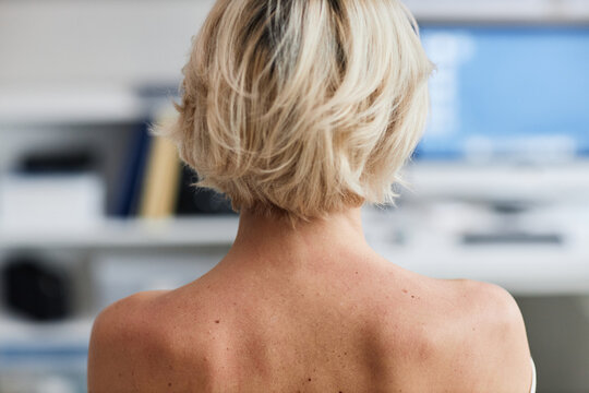 Caucasian middle aged woman sitting with bare shoulders visible from behind attending dermatologist appointment in clinical setting focusing on skin examination and consultation