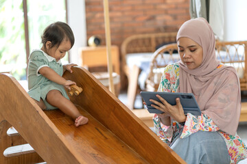 Indonesian Muslim Woman Remote Working on Tablet with Daughter Playing Indoors