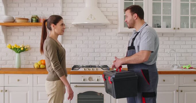 Smiling young woman shaking hands with male plumber holding toolbox after repair work in kitchen. Happy client expressing gratitude to repairman for professional help with plumbing issue at home.