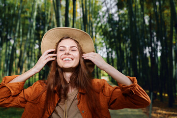 Woman smile hat bamboo forest outdoor portrait happy young in casual jacket, looking up and enjoying nature with sunlight filtering through tall green stalks in serene grove.