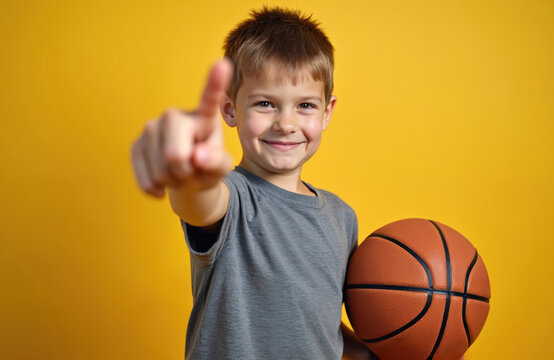Boy with basketball points finger at, inviting to play game. Child holds ball, ready for sport activity. Youngster shows confident gesture with positive facial expression against yellow backdrop. - Powered by Adobe