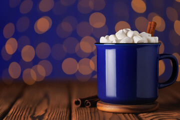 Cup of hot cocoa with marshmallows and and cinnamon sticks on wooden table against blue background of bokeh glowing garland lights