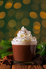 Hot chocolate with whipped cream in glass cup against green background of bokeh glowing garland lights