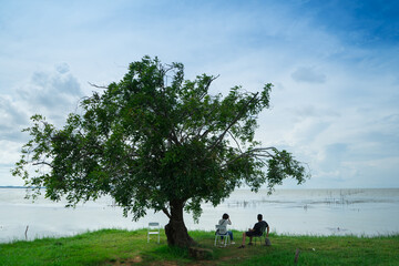 Lover sitting under the big tree near the lake .Couple tourist to relax on holiday with cloud and blue sky background on the  sunrise.