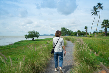 A woman to walk alone and see view of sea with along gravel walkway and flower grass at the park.