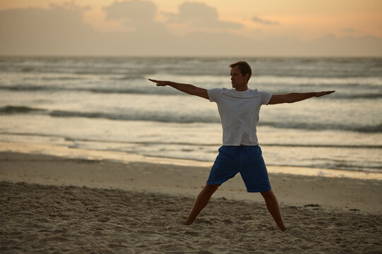 Man, stretching and exercise for yoga on beach for mindfulness, spiritual healing and zen. Person, sunset and space with warrior pose for holistic balance, inner peace and self care goals on seashore - Powered by Adobe