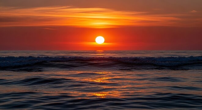 Vibrant orange sun setting over the ocean horizon, creating a beautiful solar path reflection on the dark, gentle waves under a dramatic, colorful sky