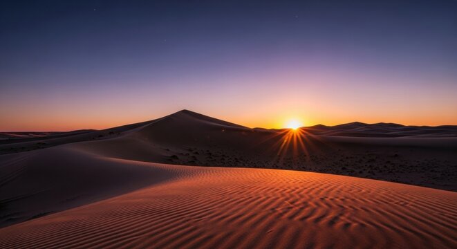 Rippled sand dunes in a vast desert landscape illuminated by the warm golden light of a setting sun, creating a dramatic sunstar effect against a clear purple and blue sky