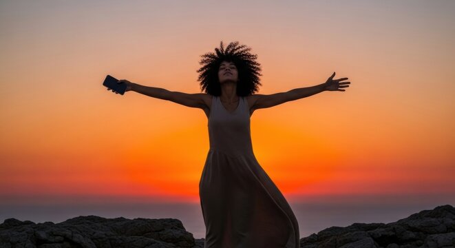 Serene woman with curly afro hair stands silhouetted with arms outstretched, holding a phone and embracing a moment of freedom against a vibrant orange sunset sky - Powered by Adobe