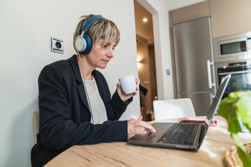 Middle aged woman with headphones sips coffee while working on laptop at kitchen table, focused on online meeting and typing