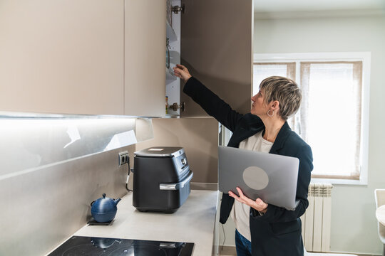 Woman managing professional tasks remotely from her modern kitchen, holding a laptop while reaching into a cabinet