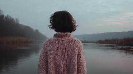 Back view of a person in a pink chunky knit sweater standing by a calm, misty river at dusk.