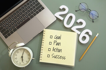flat lay of notebook with checklist of goals, plan, action and success, number of New Year 2026, pencil, laptop, alarm clock and eyeglasses on green desk. goal setting, planning for New Year concept