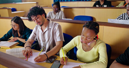 Indian young adult gen z male man sitting on desk table do study hold pencil writing exam notes college place. Focused teen pupil busy boy learn skill make new project work day time inside class room