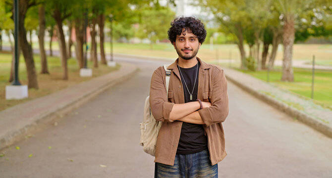 Indian happy young adult gen z man male look camera cross arm standing outdoor area. Smart cool proud pupil boy guy wear bag fold hand staring cam do casual pose enjoy new day outside place
