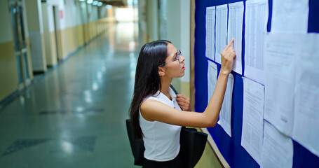 Indian young adult gen z busy female look notice board check study course exam dates results report teen girl wear bag glasses do read new day notes news paper list name data project time indoor place