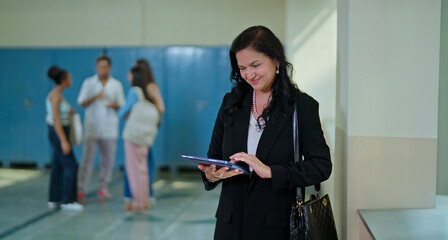 Indian college teacher standing use tablet device check online email data work task campus place. mature adult gen x happy smiling tutor lady hold tab look screen do candid casual pose inside hallway