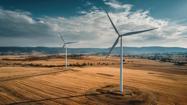 Two tall wind turbines stand in a golden farmland with a blue sky and distant hills on the horizon. Concept Wind Turbines, Golden Farmland, Blue Sky, Distant Hills, Horizon Line