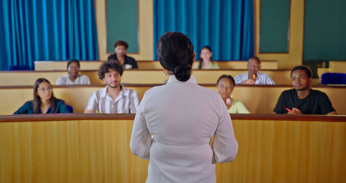 Indian woman teacher teach young adult male female sitting desk table do study learn new course exam indoor class room. Female tutor talk speak share idea plan teen group attend college day