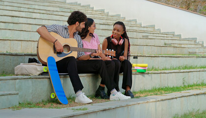 Indian young adult gen z male man hold guitar play music female sing song have fun joy outdoor place. Happy relax teen pupil girl boy group sitting on stair spend good break time enjoy new day